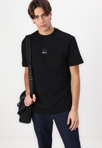 Svart kortermet t-skjorte laget av bomull, med et lite kvadratisk logo-lapp på brystet. Matchet med mørk blå denimjeans og en svart veske.
