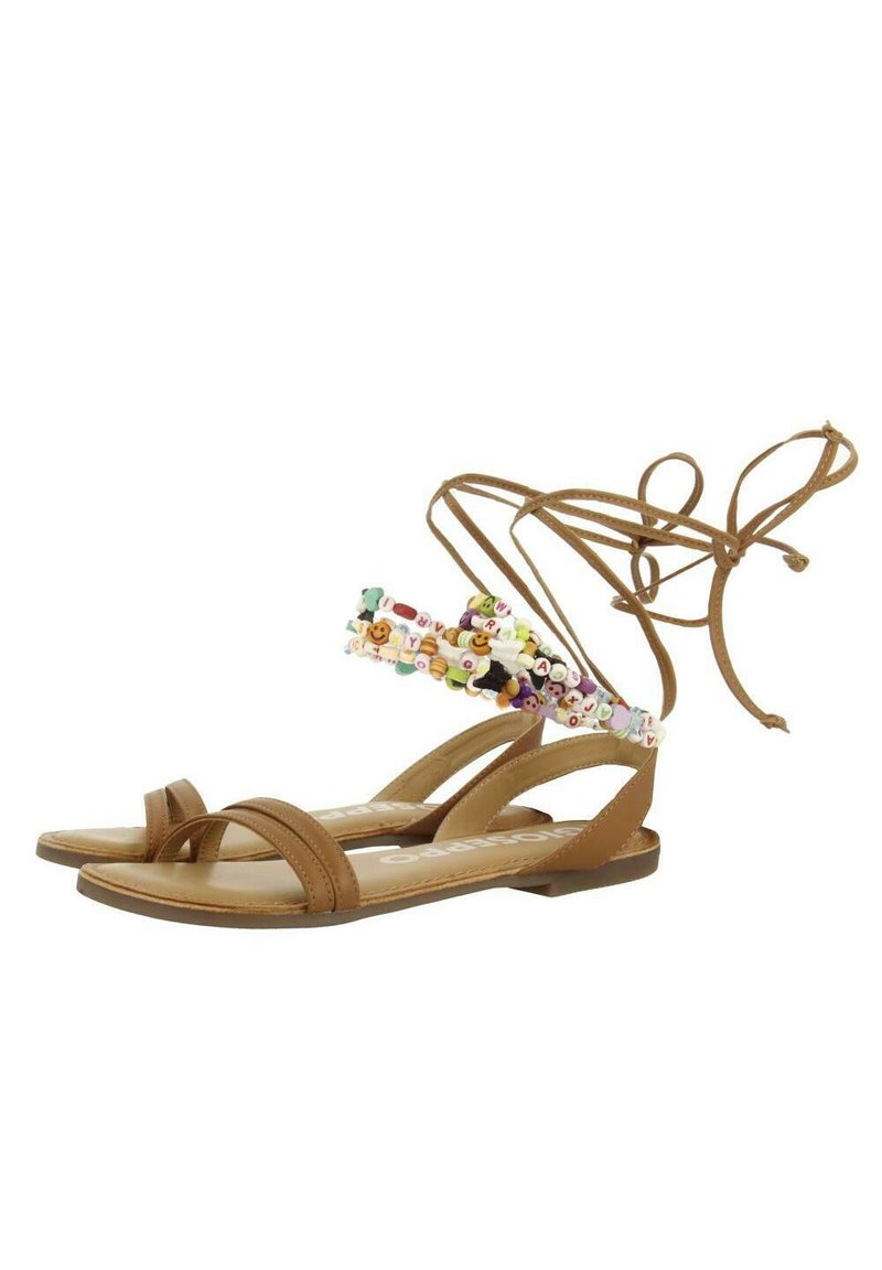 Sandalias de cuero marrón con suela plana, que presentan una colorida correa de cuentas en el tobillo y delgadas tiras entrecruzadas en los dedos.