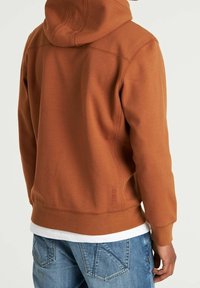 Osoba v hnědé mikině s kapucí přes bílé tričko a modré džínové kalhoty, zobrazena zezadu na jednobarevném pozadí.