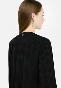 Blusa nera plissettata con scollatura rotonda e un piccolo logo sul retro. Presenta maniche lunghe e un tessuto a trama liscia.