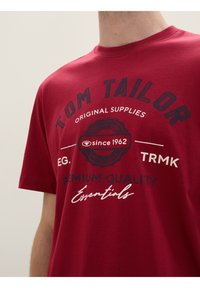 Rød bomulds T-shirt med korte ærmer, der har et cirkulært grafisk design. Teksten inkluderer "TOM TAILOR," "siden 1962," og "PRÆMIUM KVALITET."