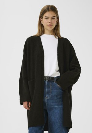 Black oversized cardigan with a soft texture, long sleeves, open front, and two side pockets, worn over a white top and blue jeans.