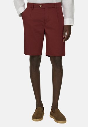 Uomo con pantaloncini bordeaux su misura, camicia bianca a maniche lunghe e espadrillas marroni senza lacci, in piedi con una mano in tasca.