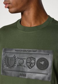 Mørkegrønn bomulls-T-skjorte med en teksturert logo-patch i svart gummi med pregete golf-relaterte grafikker og teksten "PREMIUM GOLF EST. 1997".