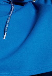 Lysblå hoodie med vita snören som har upprepade "FILA"-loggor, visar tygtextur och sömnadsdetaljer.