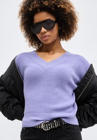 Göndör hajú nő nagy fekete napszemüveget viselve, levendulaszínű V-nyakú pulóverben, fekete szegecses övvel és fekete dzsekivel, amely éppen lecsúszik a vállairól.