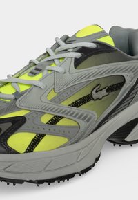 Chaussure de sport grise et jaune néon avec des accents noirs, des lacets blancs, une semelle texturée et un petit logo de reptile embossé sur le côté.