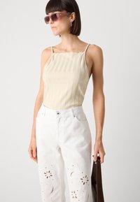 Beige rukávový top s jemnými ramínky, zkombinovaný s bílými džíny s květinovými výřezy a hnědou taškou. Celý vzhled dotvářejí hnědé sluneční brýle.