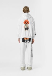 Sudadera blanca con un diseño de un perro de dibujos animados naranja en la parte posterior, combinada con pantalones blancos que tienen acentos de texto negro y un corte relajado.