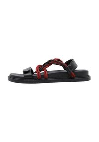 Sandalias negras con suela plana, que presentan tiras tejidas rojas que se cruzan y se envuelven por la parte superior. Material de cuero liso con un diseño minimalista.