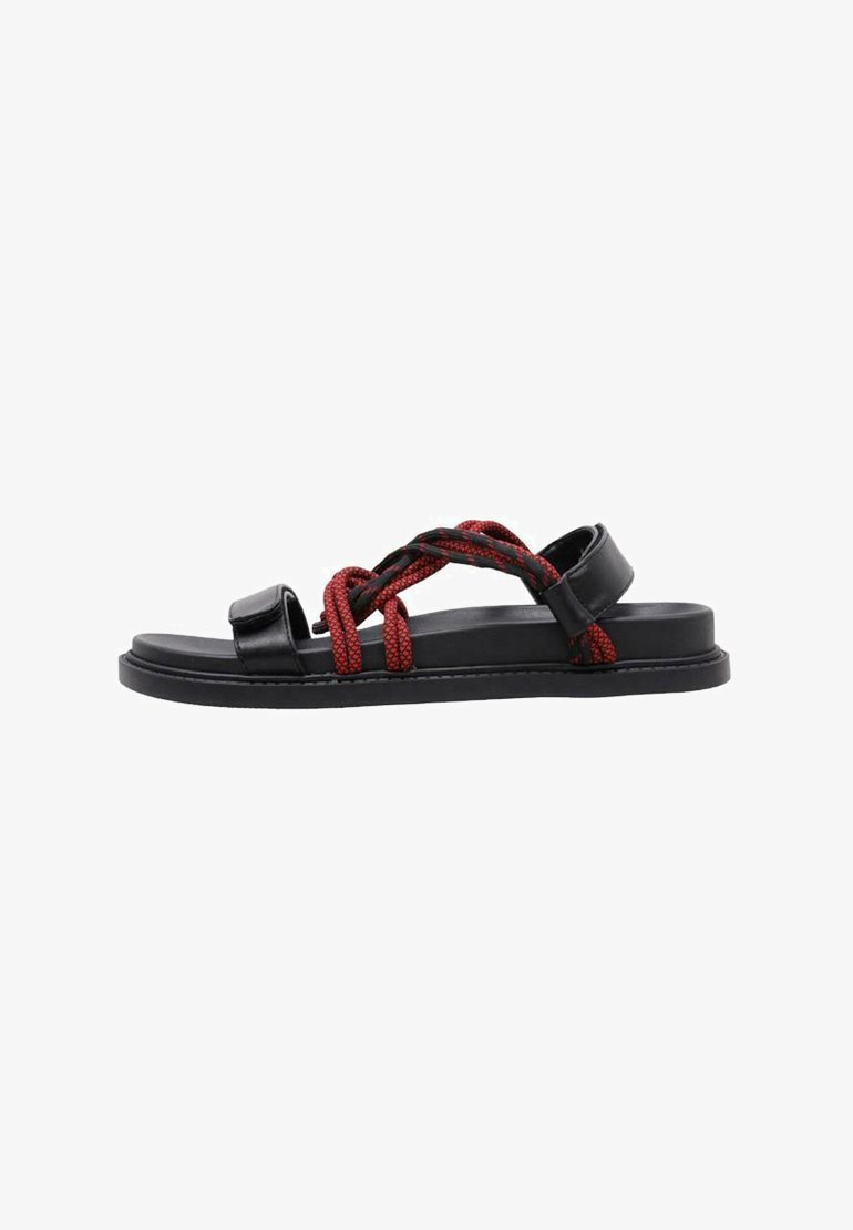 Sandalias negras con suela plana, que presentan tiras tejidas rojas que se cruzan y se envuelven por la parte superior. Material de cuero liso con un diseño minimalista.