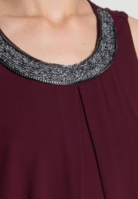 Burgundy topp med rund halsringning som har en texturerad silver- och svart kant. Slät tyg med en central veckdetalj.