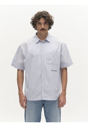 Mann mit lockigem Haar und Schnurrbart, der ein kurzärmeliges, hellblau gestreiftes Hemd mit Knöpfen und hellblaue Jeans trägt und vor weißem Hintergrund steht.