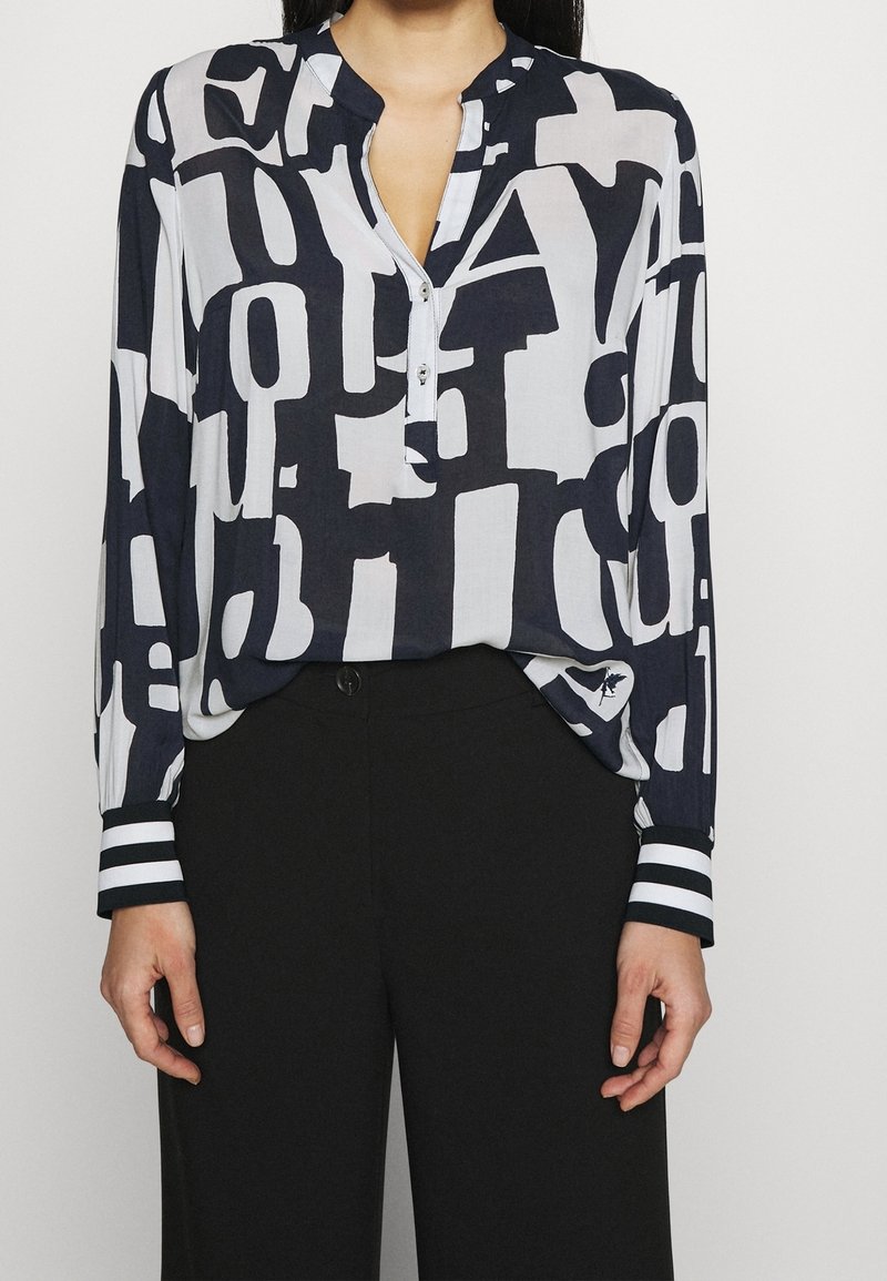 Femme portant un chemisier noir et blanc à manches longues avec des motifs de lettres abstraits et un pantalon noir, debout devant un fond uni clair.