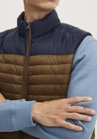 Paminkštinta liemenė su tamsiai mėlyna viršutine dalimi ir rudąja apatine dalimi, turinti aukštą apykaklę ir užtrauktuką. Po ja yra šviesiai mėlynas ilgomis rankovėmis marškinėliai.