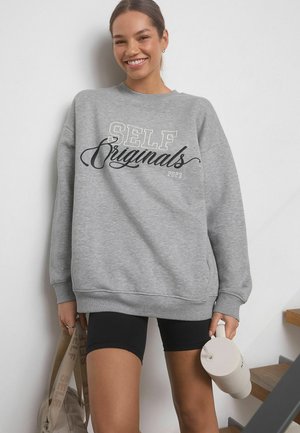 Grijze sweatshirt met "SELF Originals 2023" in zwarte en crèmekleurige tekst, gecombineerd met zwarte biker shorts en een beige waterfles.