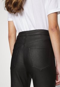 Kvinna som bär svarta byxor med hög midja och bakfickor samt en vit kortärmad skjorta instoppad, visad från midjan och nedåt.