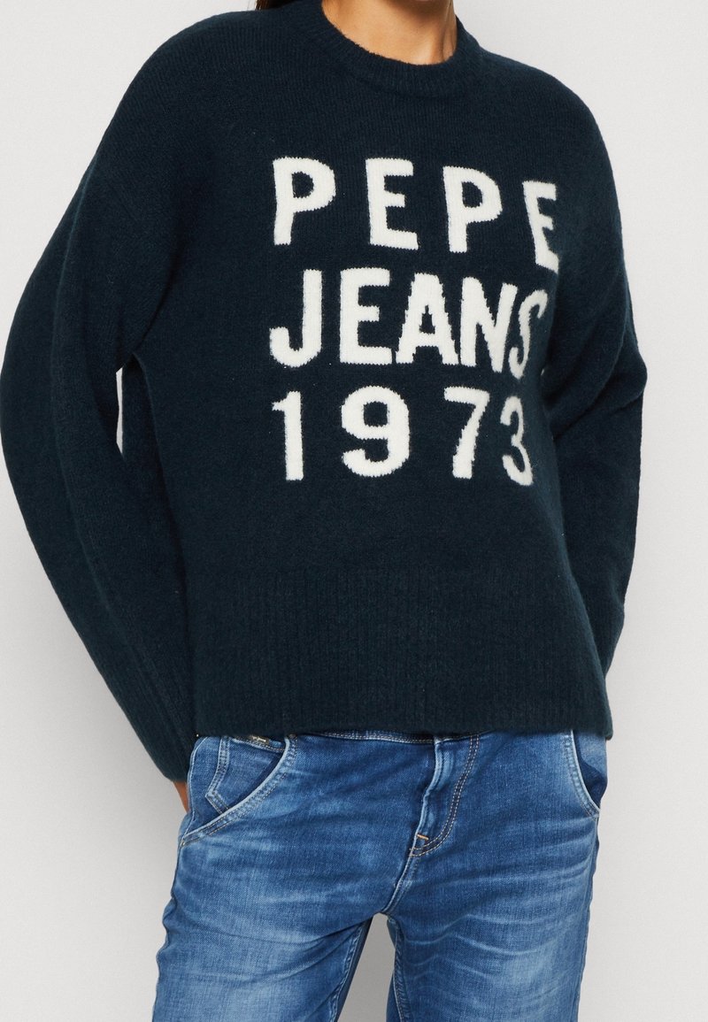 Personne portant un pull sombre avec le texte « PEPE JEANS 1973 » et un jean bleu, debout les mains dans les poches devant un fond uni.