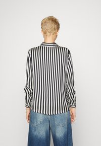 Kort blond person som står med ryggen mot oss, bär en svart och vit vertikalrandig långärmad tröja och lösa blå denimjeans.