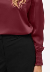Blusa di raso bordeaux con polsini arricciati, abbinata a pantaloni neri. La superficie liscia e il design aderente mettono in risalto la silhouette moderna del capo.