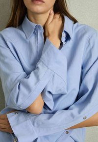 Camisa abotonada de color azul claro, con cuello, mangas sueltas y detalles de botones a presión en los puños, hecha de una tela suave con una textura lisa.