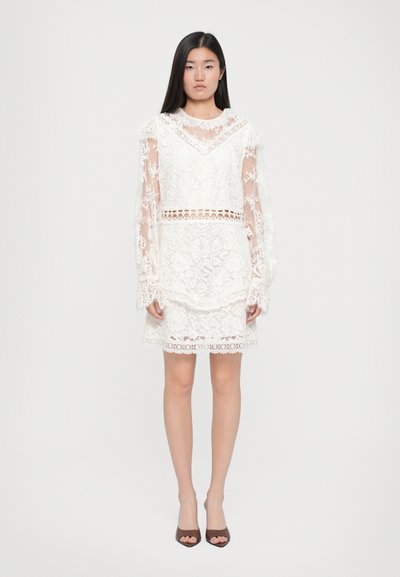 Robe en dentelle blanche avec de longues manches transparentes, des motifs floraux complexes, un corsage ajusté et un ourlet légèrement évasé. Chaussures à talons marron.