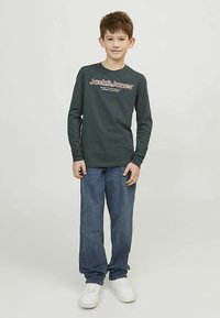 Camisa de manga larga verde oscuro con texto de logo en blanco y naranja, combinada con jeans de mezclilla azul claro y zapatillas blancas, posando sobre un fondo simple.