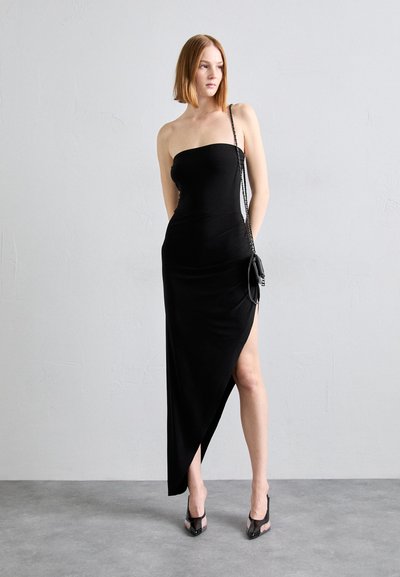 Vestito nero senza spalline realizzato in tessuto morbido, con uno spacco alto e un design aderente, abbinato a scarpe nere con tacco e una borsa piccola.