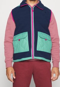 Marineblaue Fleece-Weste mit türkisfarbenen Stepptaschen und pinkem Reißverschluss. Sie verfügt über einen Kragen und zwei Fronttaschen mit Druckknopfverschluss.