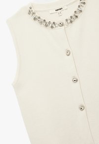 Crème mouwloos topje van een textuurstof, met een juwelenhalslijn en vier decoratieve knopen aan de voorkant.