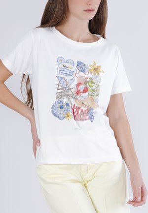 Kvinde iført hvid t-shirt med farverigt strandtema-print inklusive muslinger, solbriller, hat, søstjerne og koral, kombineret med lyse gule bukser.