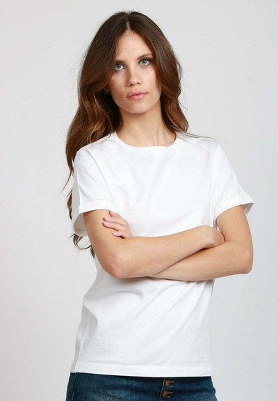 T-shirt bianca in cotone con scollatura rotonda e maniche corte, caratterizzata da una vestibilità comoda e una superficie liscia. Il modello è in piedi con le braccia incrociate.