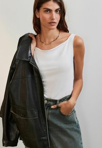 Haut blanc sans manches associé à une veste en cuir noire. Le mannequin porte une jupe en denim taille haute, mettant en valeur un collier chaîne. Textures douces et lisses.