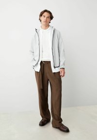 Ljusgrå lättviktsjacka med svart dragkedja, lager på lager över en vit skjorta, tillsammans med bruna byxor och mörkbruna skor.