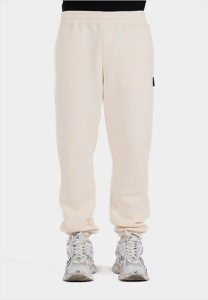 Cream-colored sweatpants made of soft fabric, feature a relaxed fit, elastic waistband, and side pockets. Paired with white sneaker shoes.