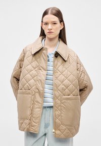 Μπεζ μπουφάν με καπιτονέ ύφασμα, μεγάλο γιακά, κουμπιά με πάτημα, δύο μπροστινές τσέπες και λογότυπο. Φορεμένο πάνω από ένα ανοιχτό μπλε ριγέ πουκάμισο.