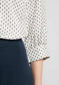 Blouse blanche avec un motif en diamants noirs, manches 3/4 et poignets retroussés, associée à une jupe texturée bleu foncé. Tissu lisse, design décontracté.