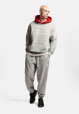 Młoda osoba z krótkimi blond włosami, ubrana w szarą bluzę z kapturem "PROSTO SPORT" z czerwoną podszewką kaptura oraz dopasowane szare spodnie dresowe, idzie z rękami w kieszeniach.