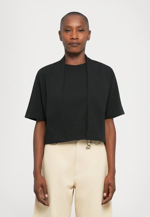 Black cropped t-shirt with short sleeves, featuring an open front design. Fabric appears soft with a smooth texture. Worn with beige pants.