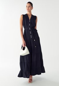 Námornícka maxi šaty s gombíkovým zapínaním vpredu, opaskom v páse, vrstveným dizajnom a volánovým lemom, skombinované s bielou štruktúrovanou kabelkou a topánkami na podpätku.
