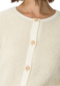 Beige Strickjacke aus weichem Stoff mit strukturierter Oberfläche, ausgestattet mit einem geraden Kragen und fünf beigen Knöpfen auf der Vorderseite.