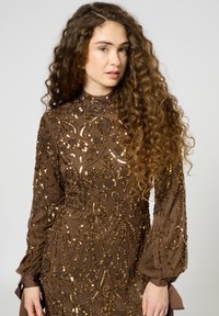 Bruine versierde jurk met ingewikkelde kraaltjes en pailletten. Lange mouwen met gerimpelde manchetten; hoge halslijn; bloemen- en spiraalpatronen.