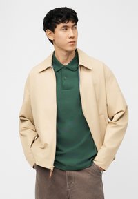 Homme aux cheveux noirs courts portant une veste beige zippée par-dessus un polo vert foncé et un pantalon marron, les mains dans les poches, regardant sur le côté.