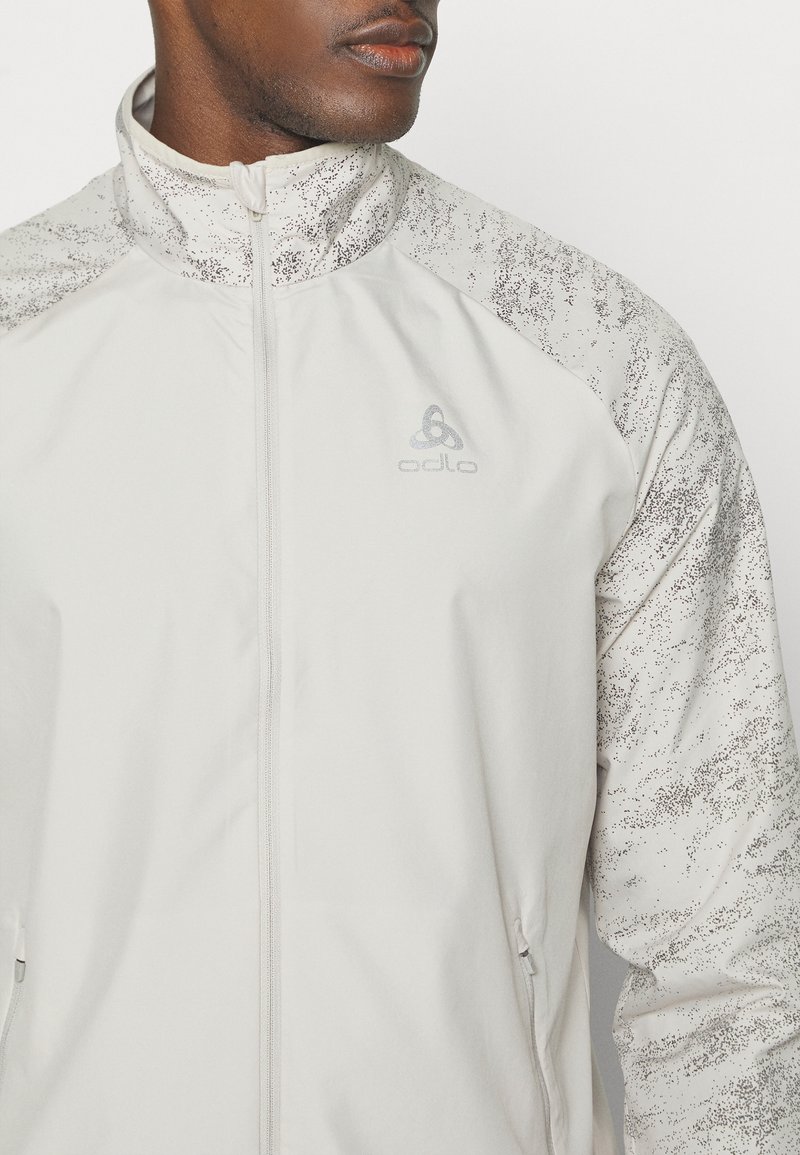 Hellgraue Sportjacke mit hohem Kragen, mit strukturierten, gemusterten Ärmeln, Reißverschlusstaschen und einem dezenten Logo auf der Brust.