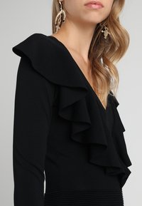 Haut noir avec un décolleté en V profond et des volants superposés aux épaules, fabriqué dans un tissu doux, avec des manches longues.