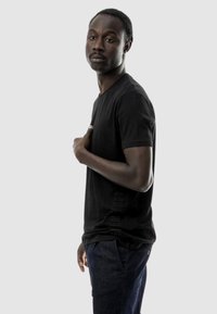 T-shirt nera a maniche corte in vestibilità rilassata, realizzata in cotone morbido con un sottile dettaglio testuale verticale sul lato. Abbinata a jeans in denim scuro.