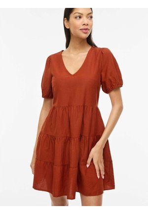 Femme portant une robe à volants couleur rouille avec des manches bouffantes et un décolleté en V, debout devant un fond blanc uni.