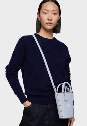 Kobieta o prostych czarnych włosach, ubrana w granatowy sweter i ciemne dżinsy, niosąca jasnoniebieską torebkę Tous z paskiem na ramię.
