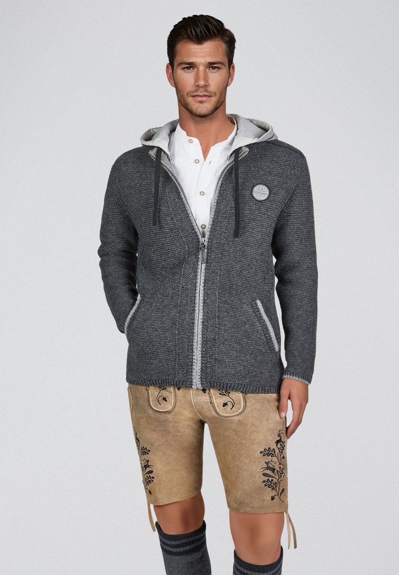 Mann trägt grauen Strick-Hoodie mit Reißverschluss, weißes Hemd, traditionelle beigen Lederhosen mit Stickerei und knielange Socken, steht vor einfachem Hintergrund.