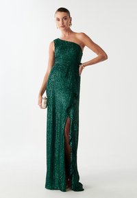 Groene paillettenjurk met één schouderontwerp, getailleerde silhuet, zijsplit en ruches. Materiaal heeft een glanzende, gestructureerde afwerking.